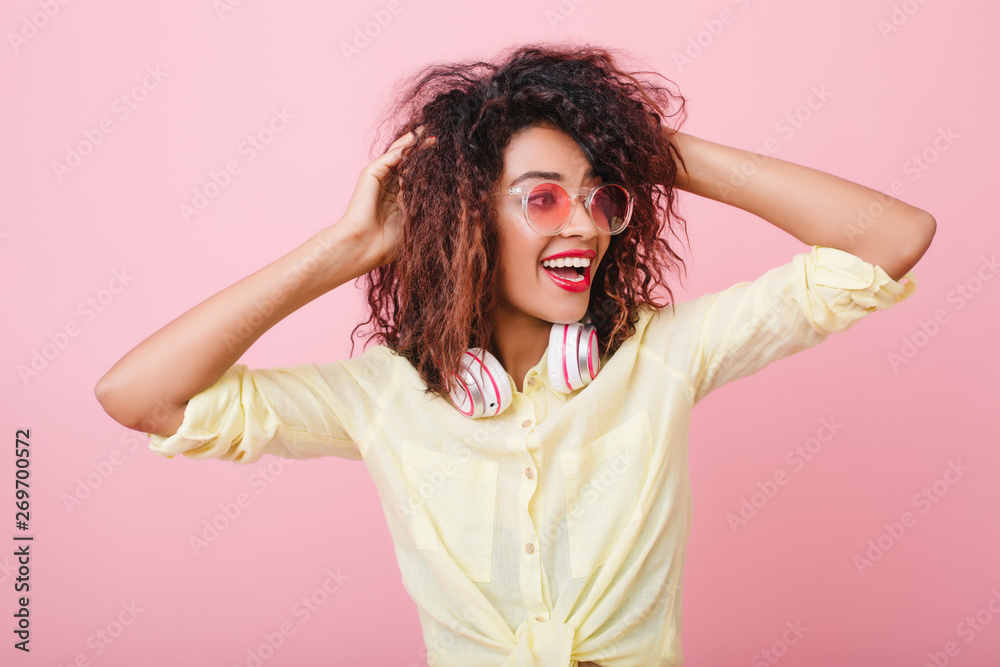 Romantic girl in cute yellow shirt enjoying fashion phooshoot in studio with bright interior. Lovable black lady in sunglasses touching her curly hair with pleasure. Romantic girl in cute yellow shirt enjoying fashion phooshoot in studio with bright interior. Lovable black lady in sunglasses touching her curly hair with pleasure.