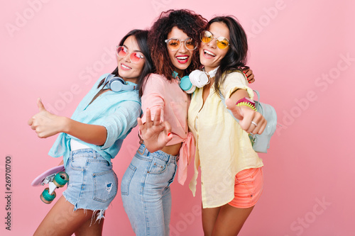 Three international female friends in headphones happy dancing on pink background with thums up. Indoor portrait of slim asian girl holding skateboard and posing with african and latin ladies. photo