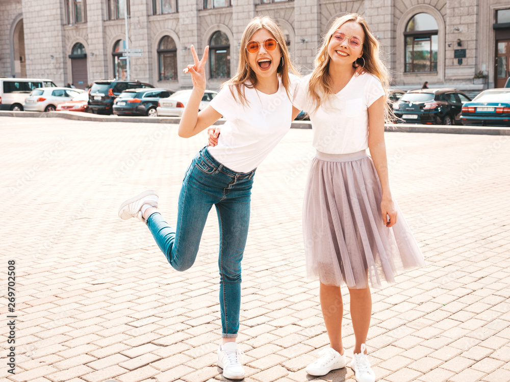 Portrait of two young beautiful blond smiling hipster girls in trendy summer white t-shirt clothes. Sexy carefree women posing on street background. Positive models having fun in sunglasses.Hugging