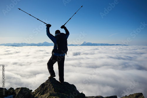 Back view of tourist hiker with backpack standing on top of huge rock with trekking poles in raised arms on bright blue sky at dawn, foggy valley filled with white clouds and mountain tops background.