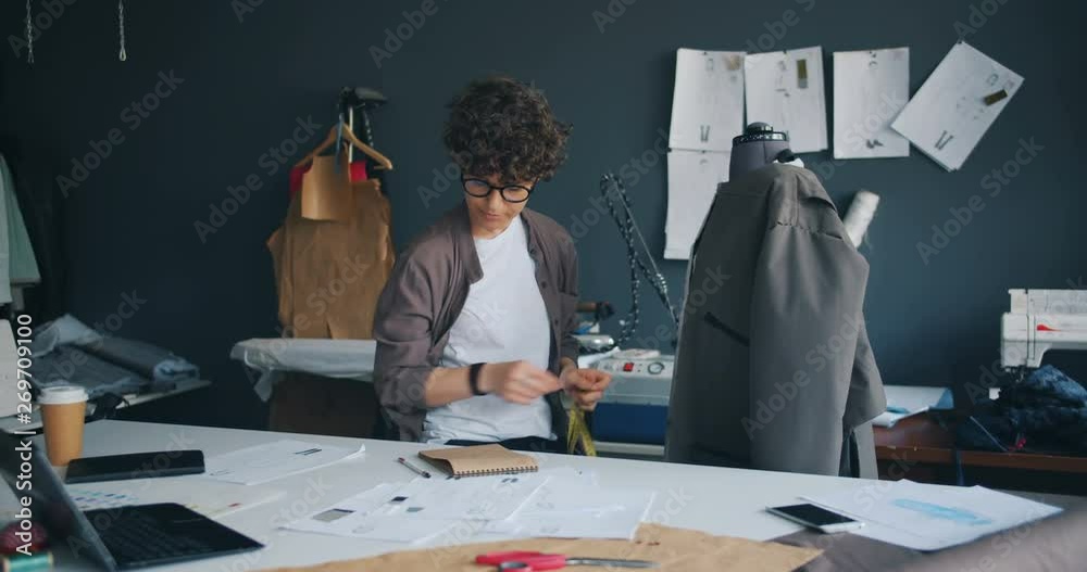 Young woman clothes designer measuring coat with measure-tape writing notes in notebook concentrated on process working in modern stydio. People and job concept.