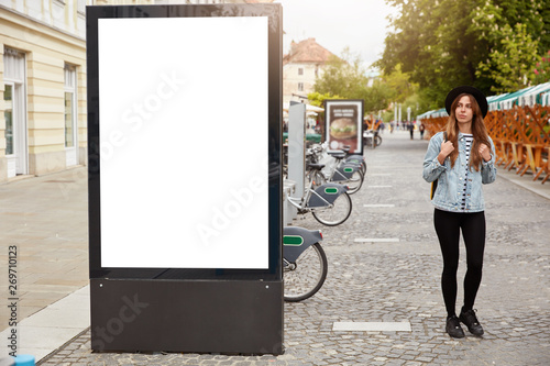 Thoughtful female tourist strolls on footway near lightbox with mock up blank space for your advertising content or commercial information. Street style concept. Focus on billboard at sidewalk