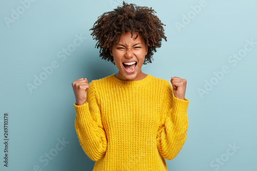 Successful dark skinned female student happy to get scholarship, clenches fists, accomplishes goal, exclaims finally victory, stands amused over blue background. Success, cheer and achievement concept