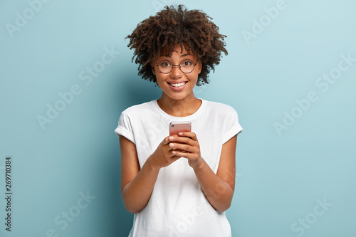 Half length shot of Afro woman holds mobile phone, enjoys nice talk online in social networks, reads funny article in internet, uses cellular, wears white t shirt and spectacles, isolated on blue wall