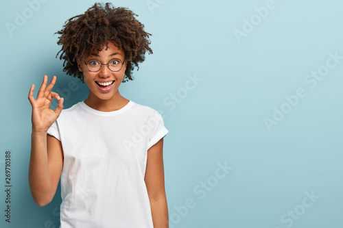 Horizontal shot of cheerful Afro American woman makes okay gesture, agrees with good proposal, wears spectacles and white casual t shirt, says incredible offer, isolated over blue background photo
