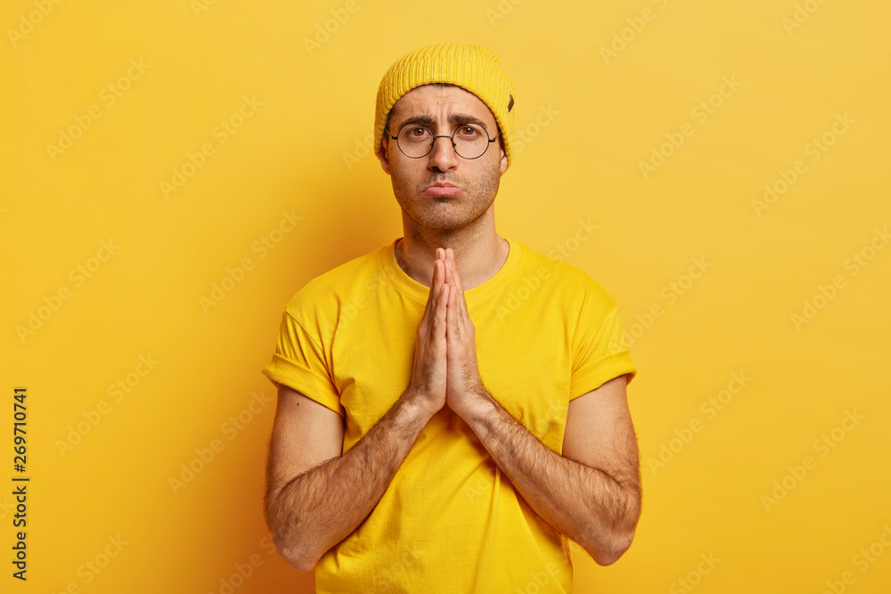 Sad male model with pleading begging expression, keeps palms together, feels guilty, asks for forgiveness, wears round spectacles, dressed in hat and t shirt, isolated over yellow background Sad male model with pleading begging expression, keeps palms together, feels guilty, asks for forgiveness, wears round spectacles, dressed in hat and t shirt, isolated over yellow background