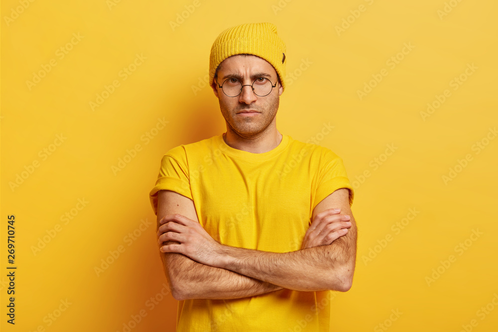 Portrait of dissatisfied young man with dark eyes, stubble, keeps arms folded, smirks face in dissatisfaction, wears yellow hat and t shirt, being angry with someone. Negative facial expressions