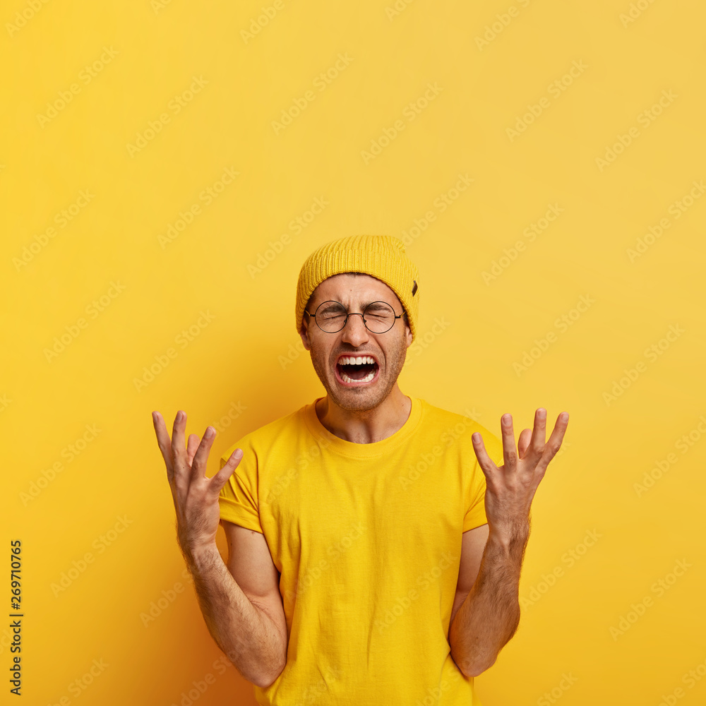 Devastated stressful sorowful man cries and gestures angrily, regrets bad mistake, has defeat, shouts from negative emotions, wears yellow hat casual t shirt, stands on yellow studio wall, blank space