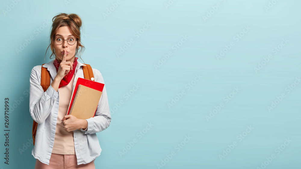 Surprised female student makes shush gesture, looks in embarrassement, holds notebooks, gossips with groupmates, tells secret information, stands indoor over blue studio wall with empty space Surprised female student makes shush gesture, looks in embarrassement, holds notebooks, gossips with groupmates, tells secret information, stands indoor over blue studio wall with empty space