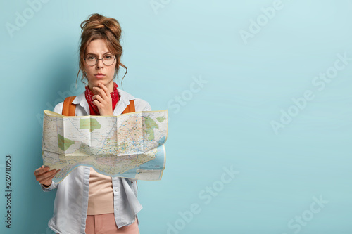 Studio shot of serious thoughtful female tourist holds chin, plans her trip, holds paper map, wears transparent glasses, looks straightly at camera, stands against blue background with free space