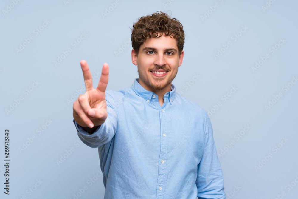 Blonde man over blue wall smiling and showing victory sign Blonde man over blue wall smiling and showing victory sign