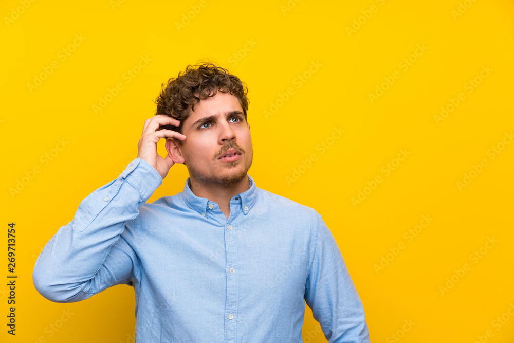 Blonde man over isolated yellow wall having doubts and with confuse face expression Blonde man over isolated yellow wall having doubts and with confuse face expression