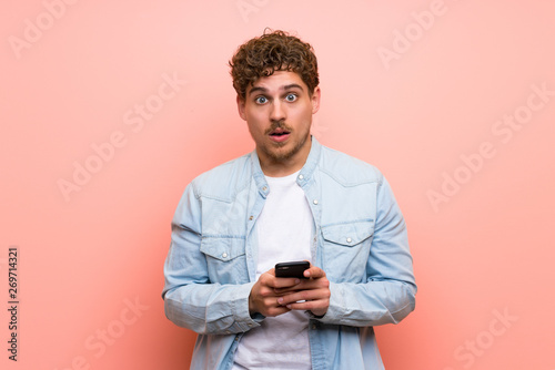 Blonde man over pink wall surprised and sending a message