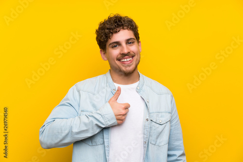 Blonde man over isolated yellow wall giving a thumbs up gesture