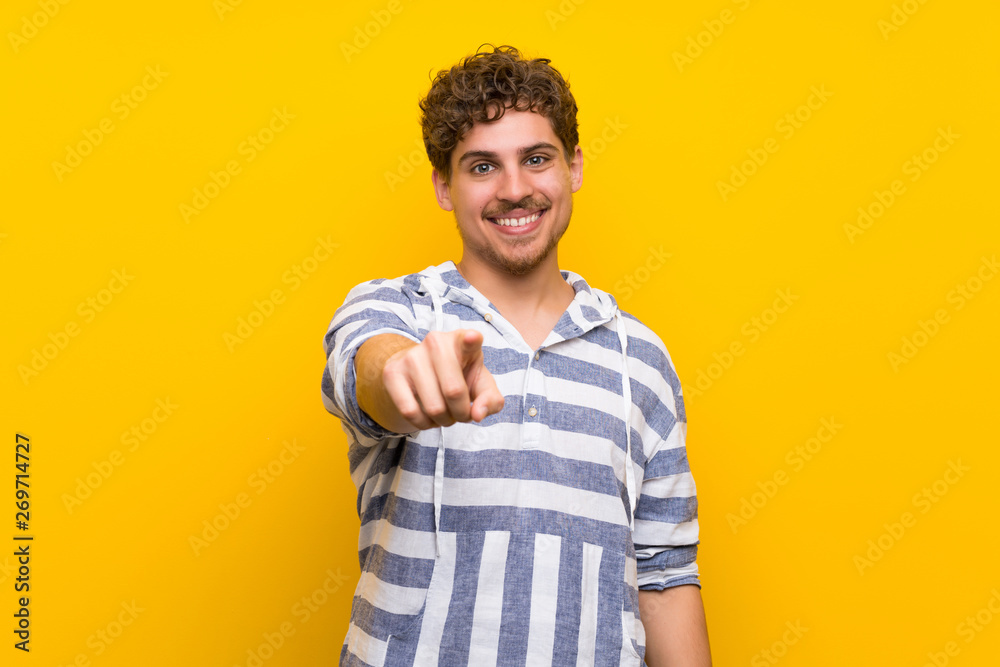 Blonde man over yellow wall points finger at you with a confident expression Blonde man over yellow wall points finger at you with a confident expression