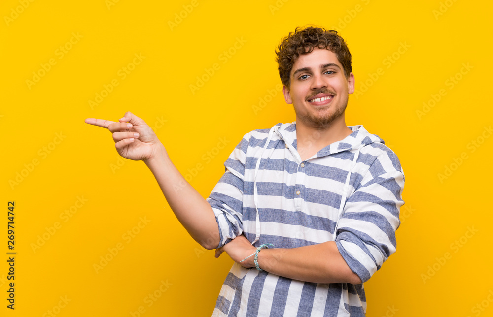 Blonde man over yellow wall pointing finger to the side in lateral position Blonde man over yellow wall pointing finger to the side in lateral position