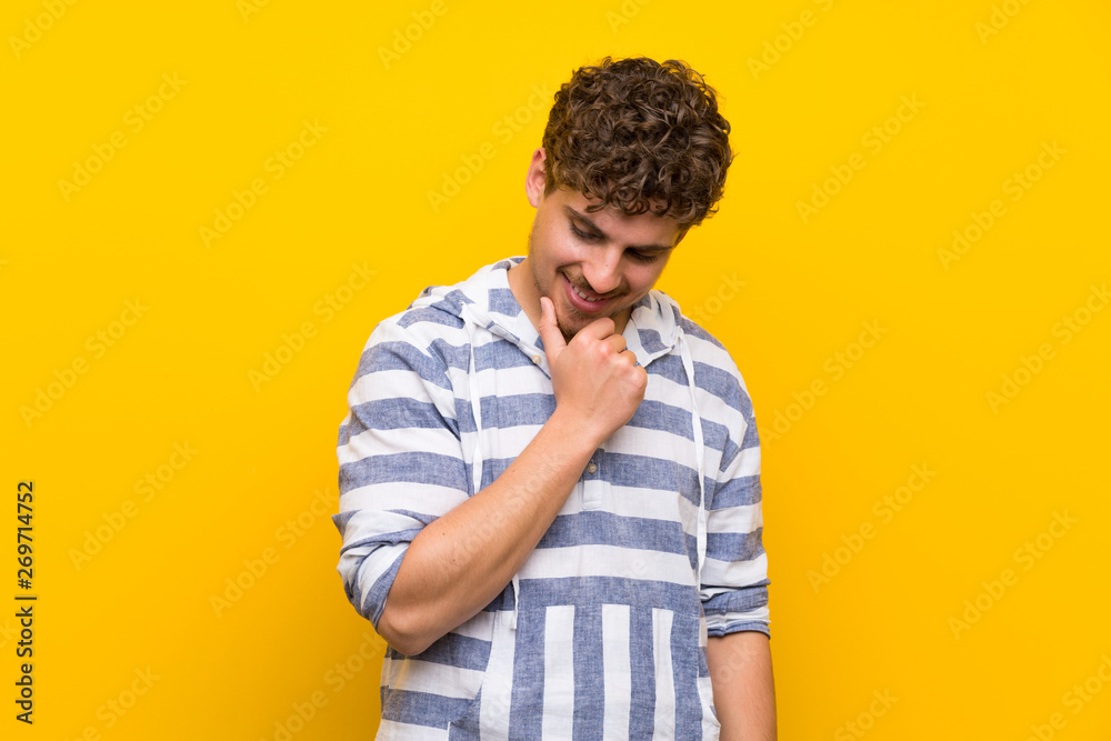 Blonde man over yellow wall looking down with the hand on the chin Blonde man over yellow wall looking down with the hand on the chin