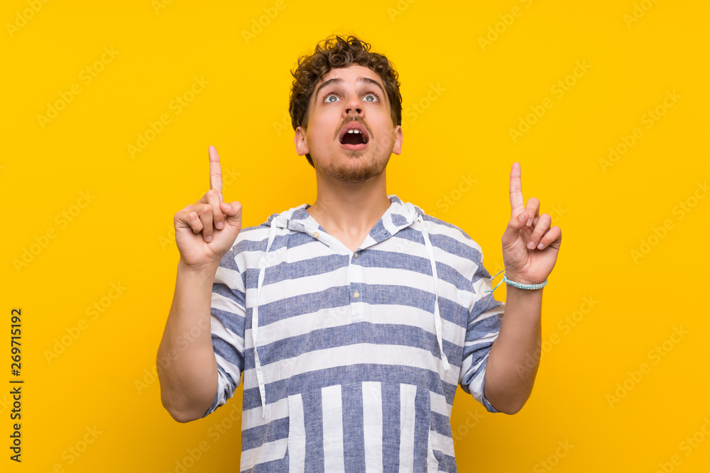 Blonde man over yellow wall surprised and pointing up