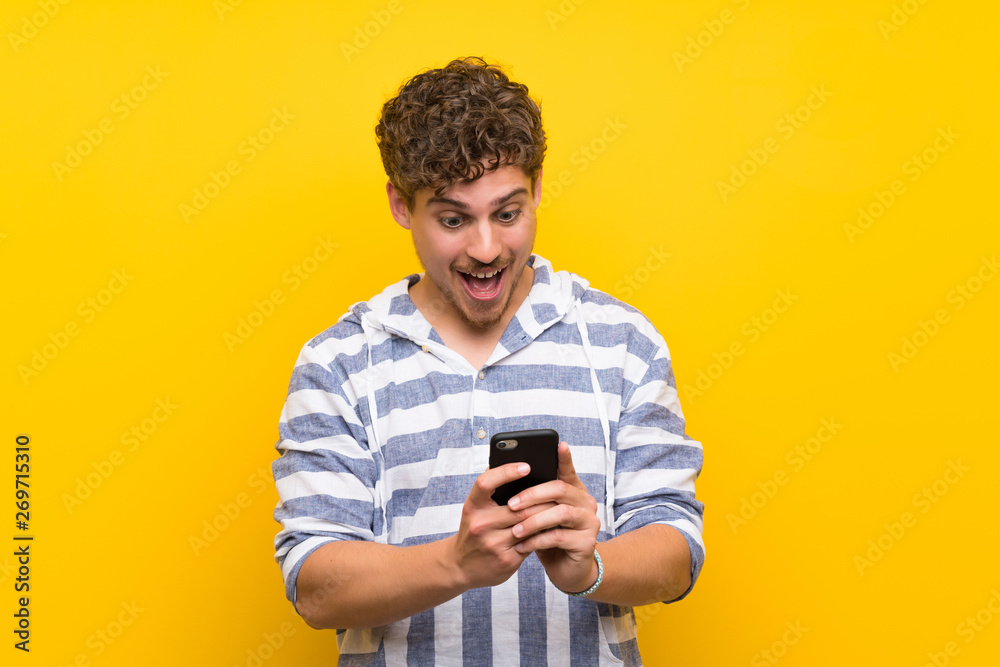 Blonde man over yellow wall surprised with a mobile Blonde man over yellow wall surprised with a mobile