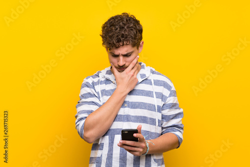 Blonde man over yellow wall with a mobile and thinking