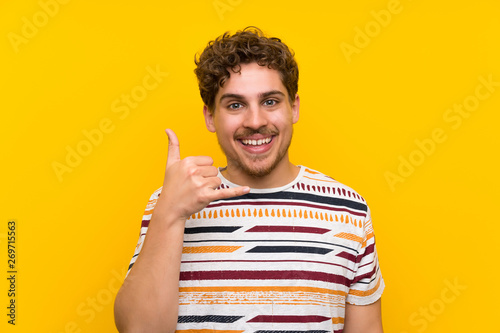 Blonde man over yellow wall making phone gesture