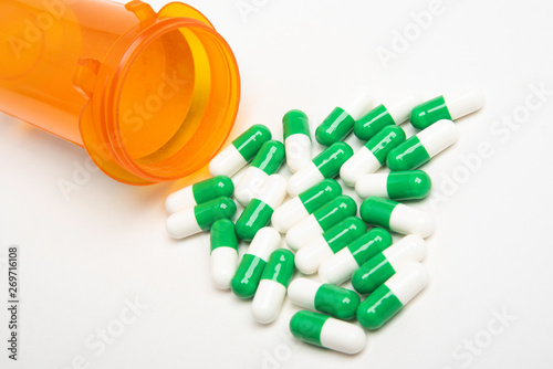 An open and capless orange plastic medicine bottle with white and green pills spilling out and into a plain white background.