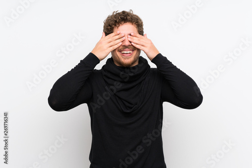 Blonde man over isolated white wall covering eyes by hands