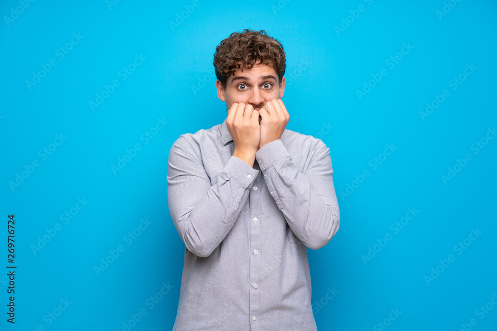 Blonde man over blue wall nervous and scared putting hands to mouth Blonde man over blue wall nervous and scared putting hands to mouth