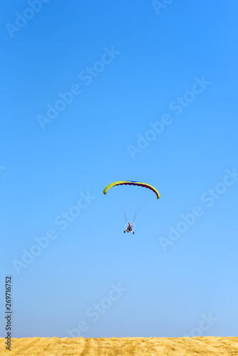 Powered paraglider parachute against blue sky and yellow field. Paramotor flying in sunny day. Freedom and summer holiday concept, extreme sport. 