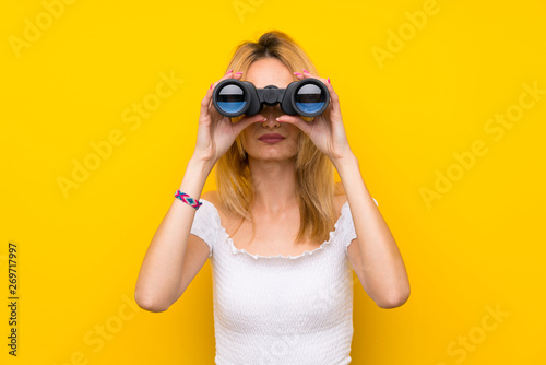 Young blonde woman over isolated yellow wall with black binoculars