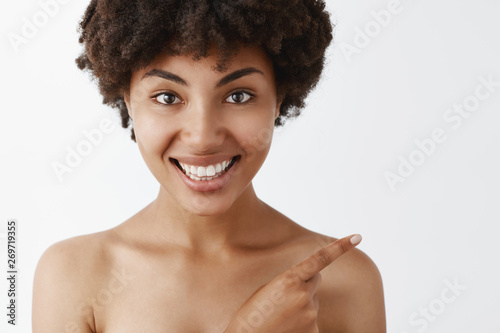 Close-up shot of feminine attractive and beautiful naked dark-skinned woman with curly hairstyle pointing at upper right corner and smiling joyfully helping out to find way