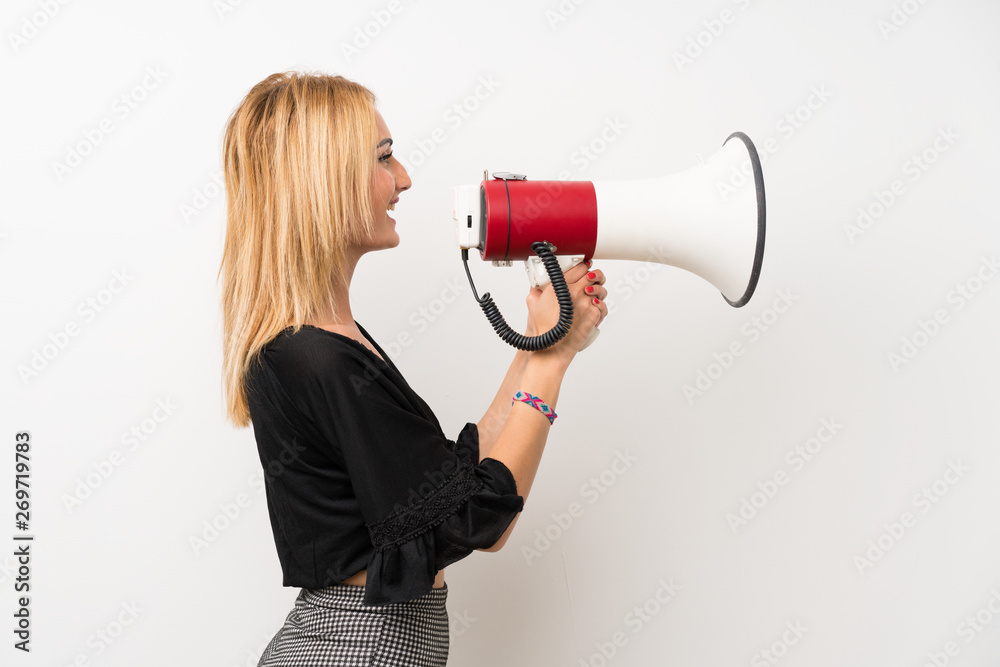 Young blonde woman over isolated white wall shouting through a megaphone Young blonde woman over isolated white wall shouting through a megaphone