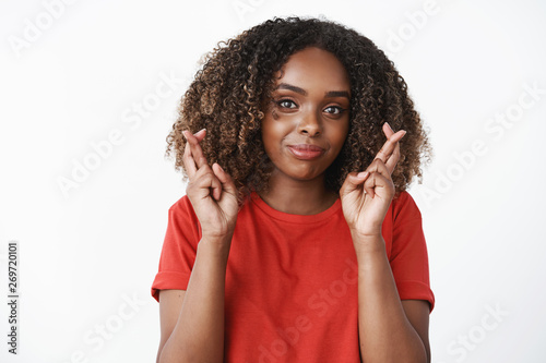 Cross my fingers for our goal. Confident good-looking optimistic african american female in red t-shirt having faith everything be alright making wish and praying smiling at camera over white wall © Cookie Studio