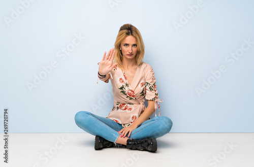 Young blonde woman sitting on the floor making stop gesture