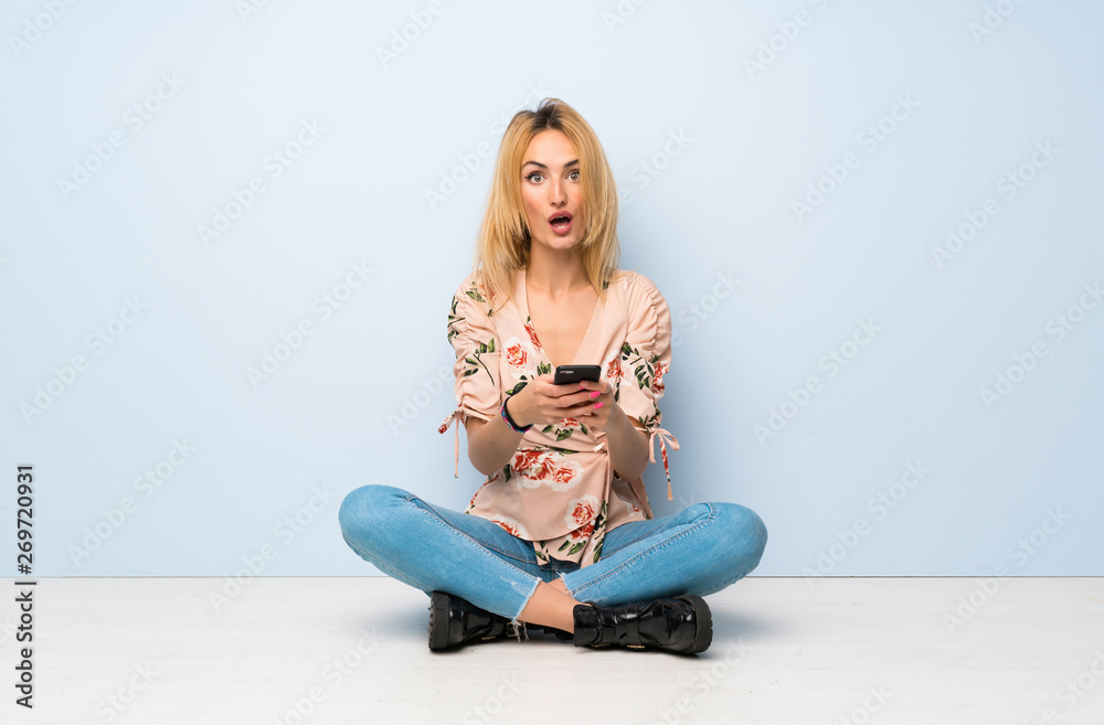 Young blonde woman sitting on the floor surprised and sending a message Young blonde woman sitting on the floor surprised and sending a message