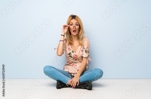 Young blonde woman sitting on the floor with glasses and surprised