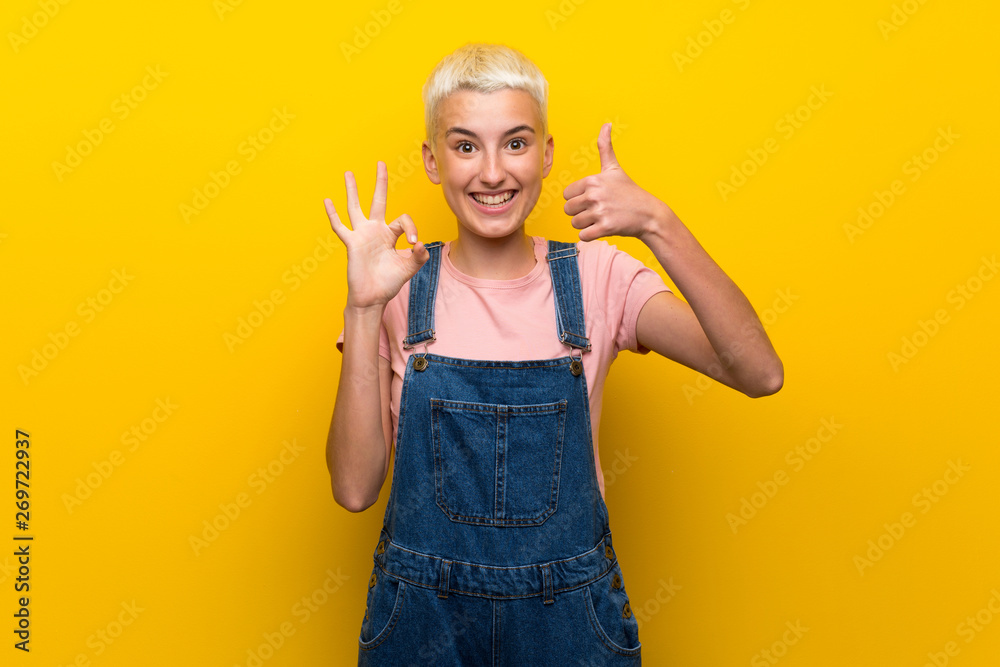 Teenager girl with overalls on yellow background showing ok sign and thumb up gesture Teenager girl with overalls on yellow background showing ok sign and thumb up gesture