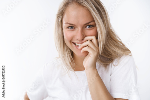 Gentle and tender young attractive blond girlfriend giggling flirty covering smile with fingers gazing at camera playfully, laughing staying in good mood look after skin condition over white wall