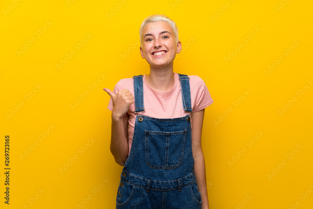 Teenager girl with overalls on yellow background pointing to the side to present a product Teenager girl with overalls on yellow background pointing to the side to present a product
