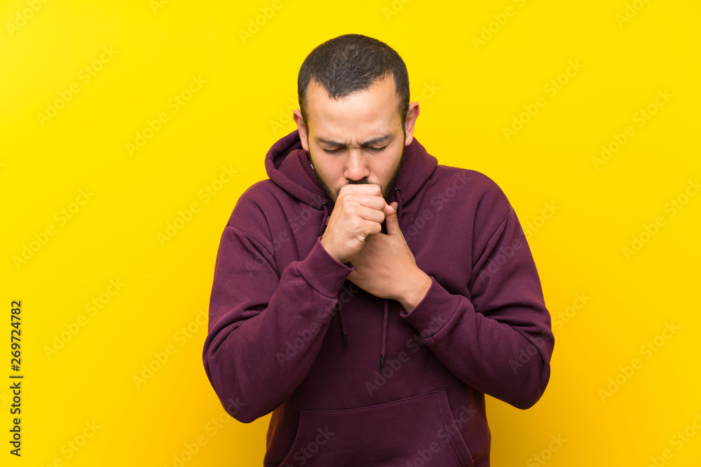 Colombian man with sweatshirt over yellow wall is suffering with cough and feeling bad Colombian man with sweatshirt over yellow wall is suffering with cough and feeling bad