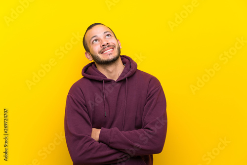 Colombian man with sweatshirt over yellow wall looking up while smiling © luismolinero