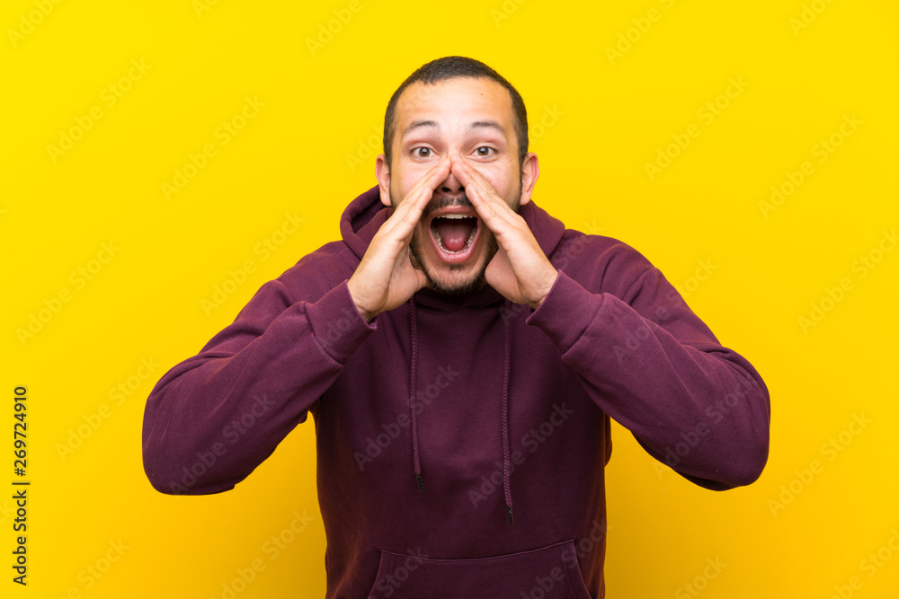 Colombian man with sweatshirt over yellow wall shouting and announcing something