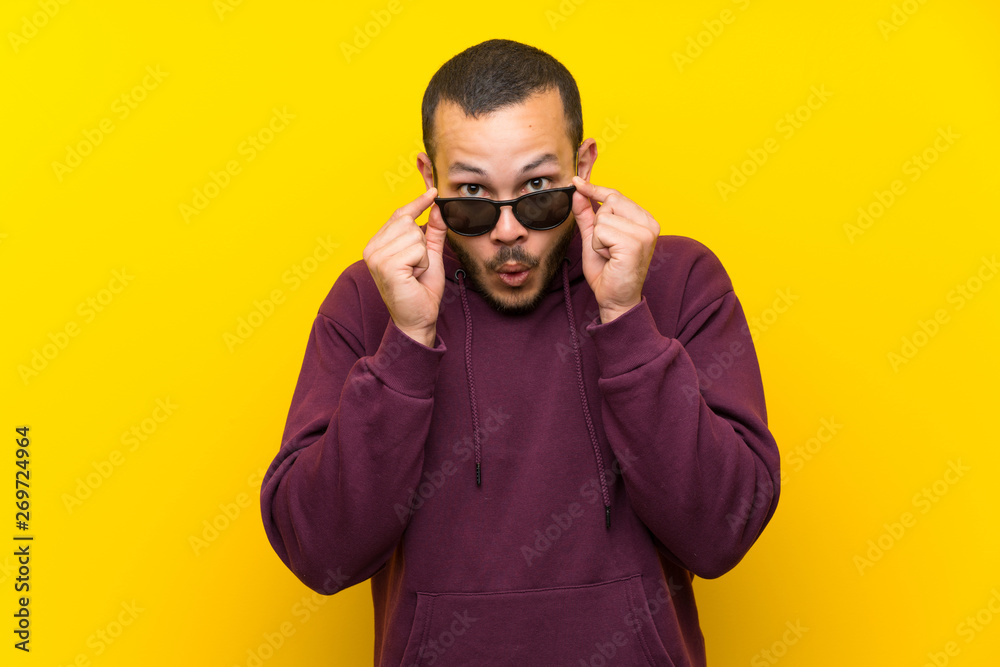 Colombian man with sweatshirt over yellow wall with glasses and surprised Colombian man with sweatshirt over yellow wall with glasses and surprised