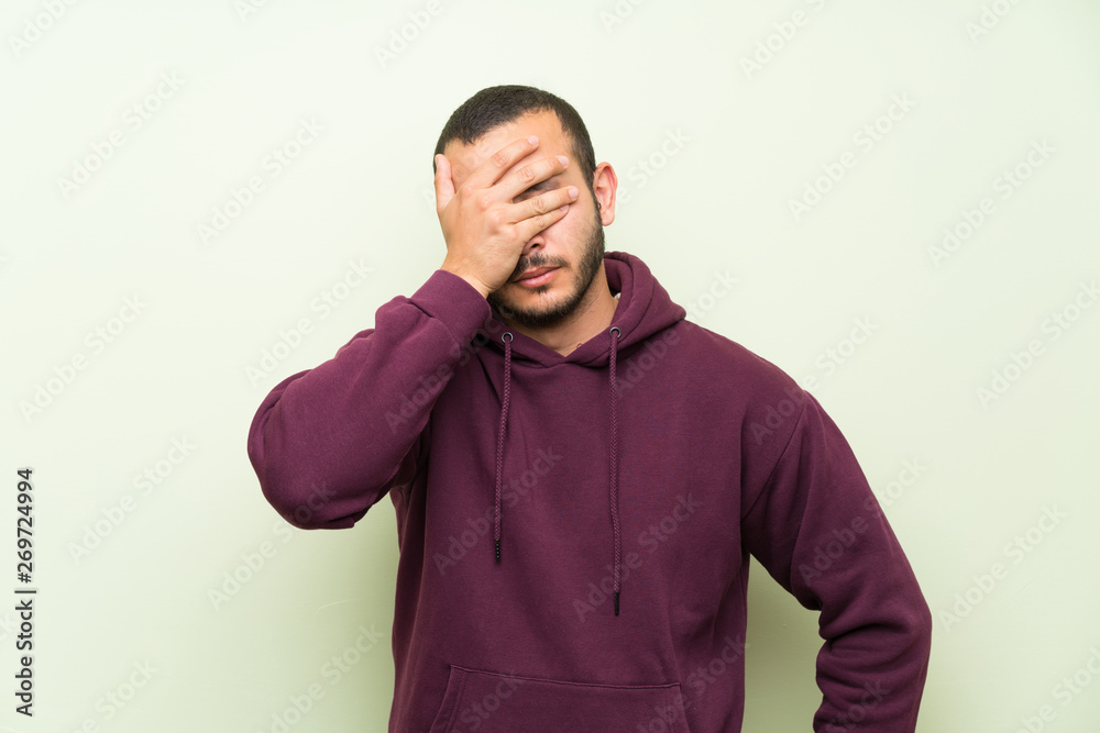 Colombian man with sweatshirt over green wall covering eyes by hands