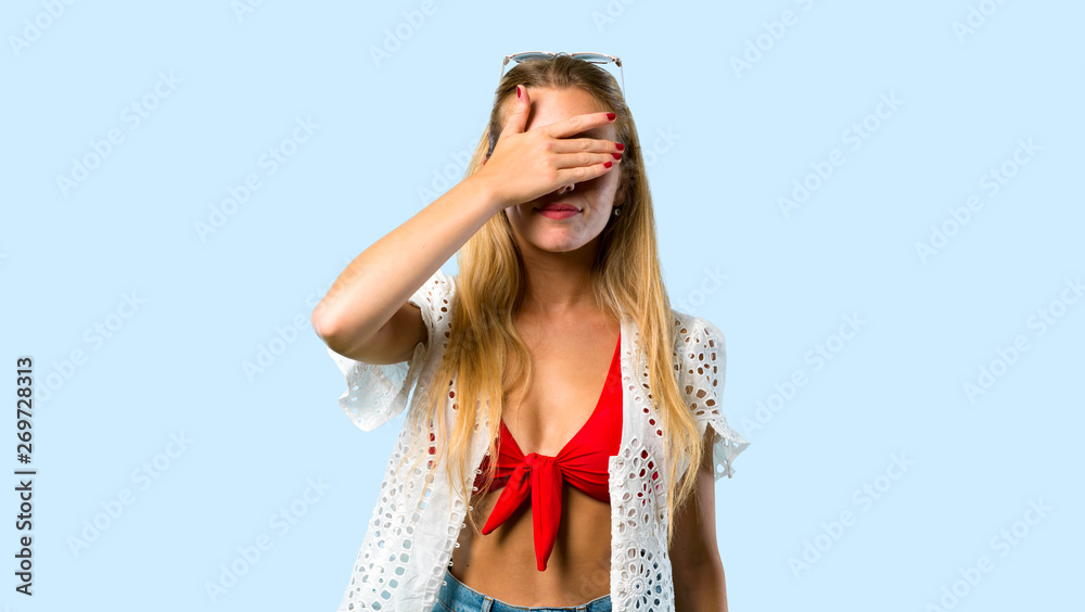 Blonde girl in summer vacation covering eyes by hands. Do not want to see something on blue background