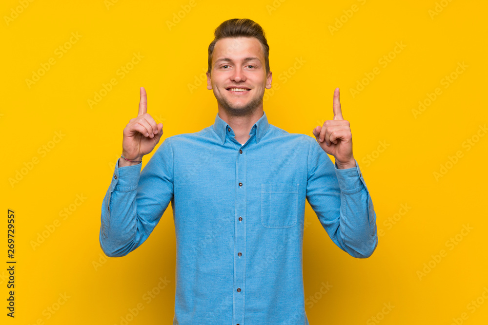 Blonde man over isolated yellow wall pointing up a great idea Blonde man over isolated yellow wall pointing up a great idea