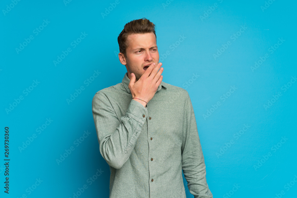 Blonde man over isolated blue wall yawning and covering wide open mouth with hand