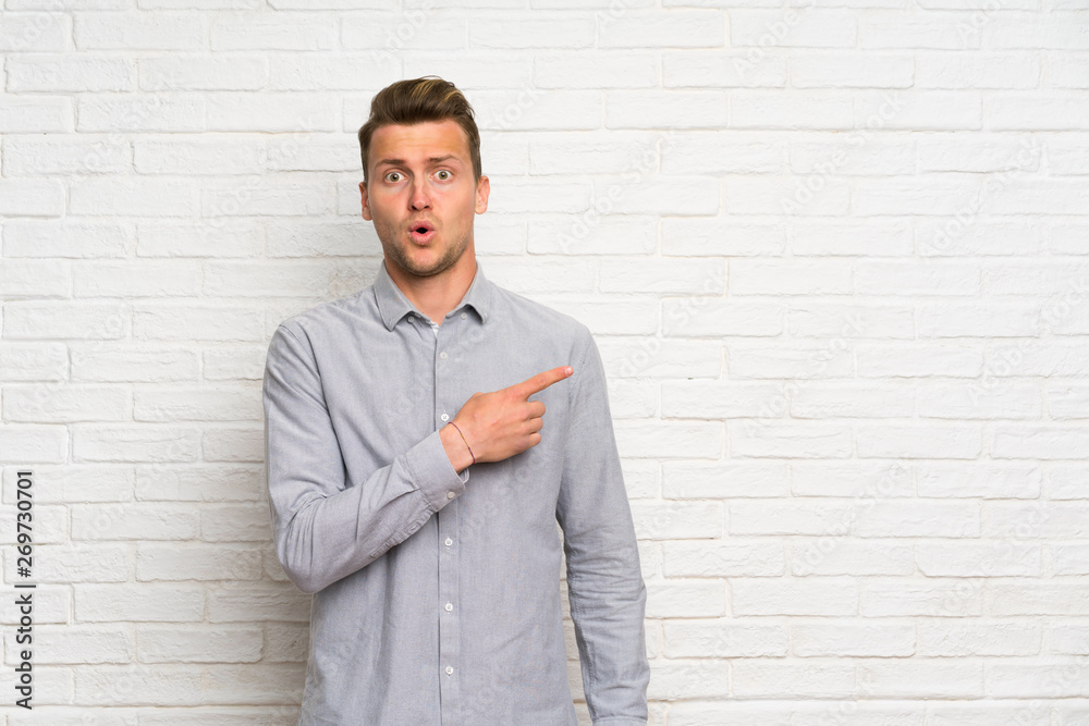 Blonde man over white brick wall surprised and pointing side Blonde man over white brick wall surprised and pointing side