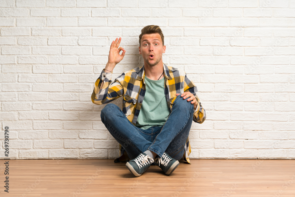 Blonde man sitting on the floor surprised and showing ok sign Blonde man sitting on the floor surprised and showing ok sign