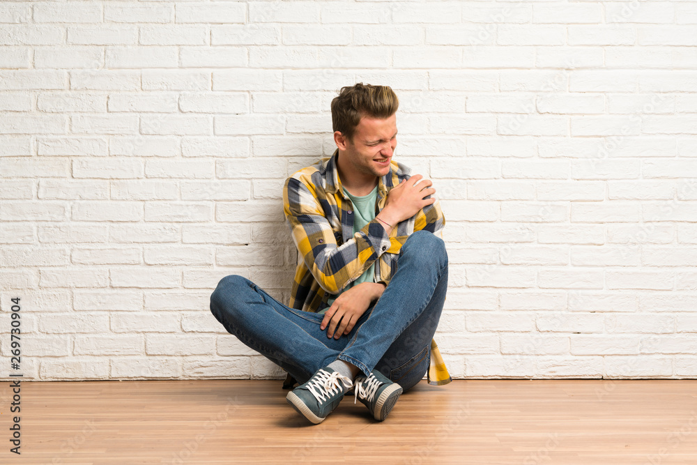 Blonde man sitting on the floor suffering from pain in shoulder for having made an effort Blonde man sitting on the floor suffering from pain in shoulder for having made an effort